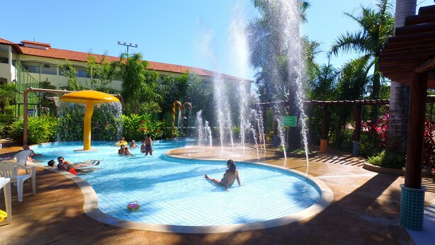 A swimming pool area with multiple water features, including fountains and a water slide, surrounded by lush greenery and a building with a red-tiled roof. - Agape - Hot Springs Apart Hotel (Caldas Novas)