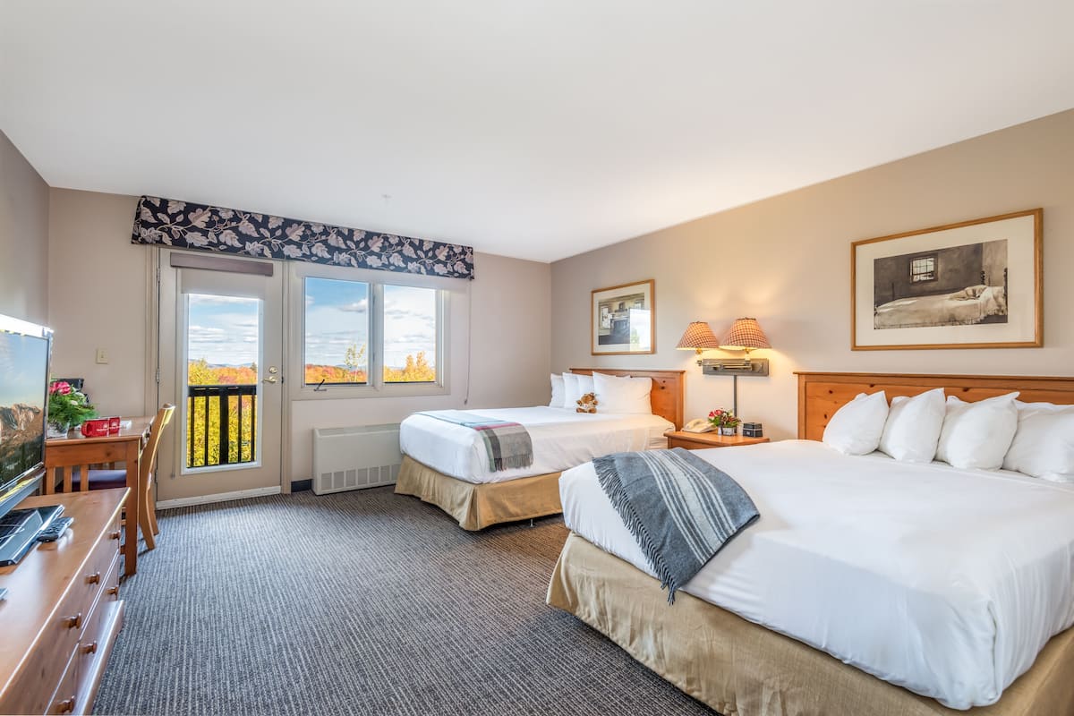 Quarto de hotel com duas camas, uma escrivaninha, uma cadeira, televisão e varanda com vista para árvores de outono.