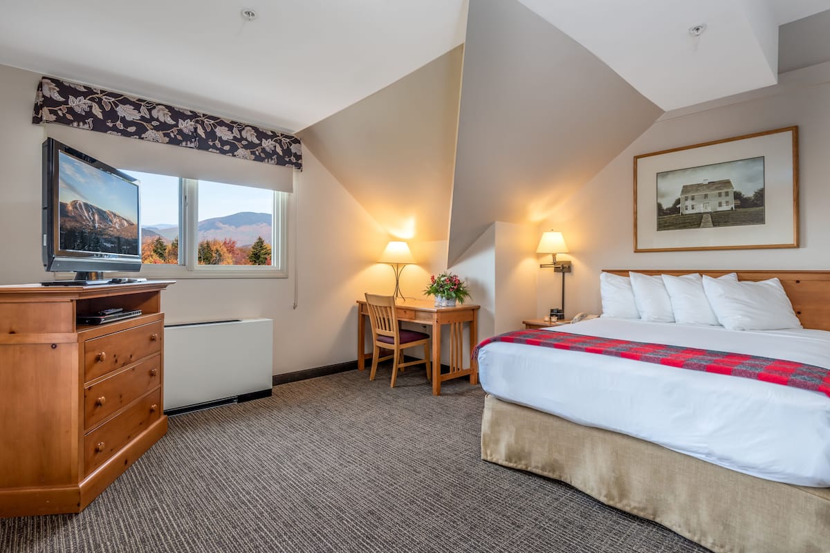 Quarto de hotel com cama, escrivaninha, cadeira, televisão e janela com vista para montanhas.