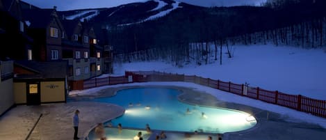 A heated outdoor swimming pool surrounded by snow, with a mountain in the background.
