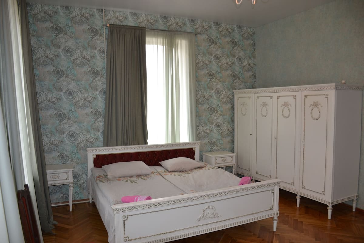 Un dormitorio con cama, armario, mesita de noche y ventana con cortinas.
