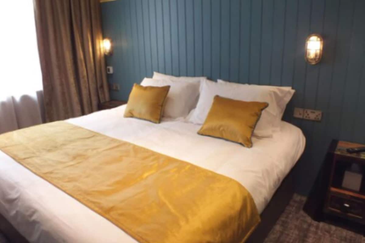 Quarto de hotel com uma cama grande, duas almofadas, um cobertor dourado e uma mesa de cabeceira com abajur.