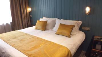 A hotel room with a large bed, two pillows, a gold throw blanket, and a nightstand with a lamp.
