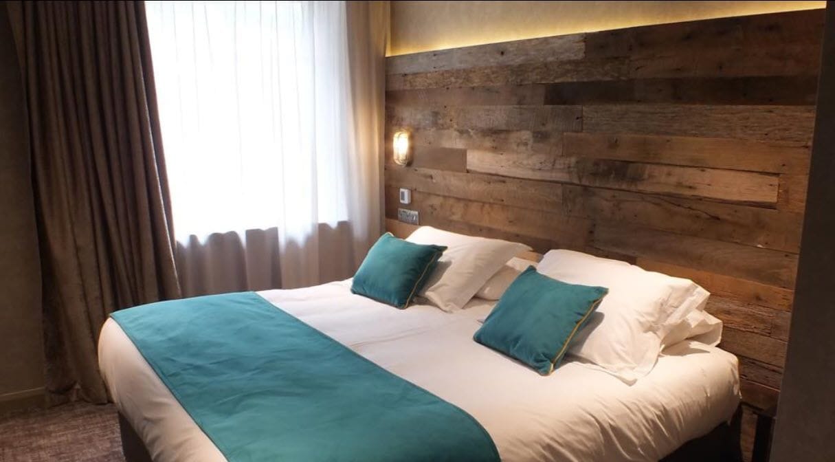 Una habitación de hotel con cabecera de madera, una cama grande con ropa de cama azul verdosa y blanca, y una ventana con cortinas.