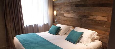Una habitación de hotel con cabecera de madera, una cama grande con ropa de cama azul verdosa y blanca, y una ventana con cortinas.