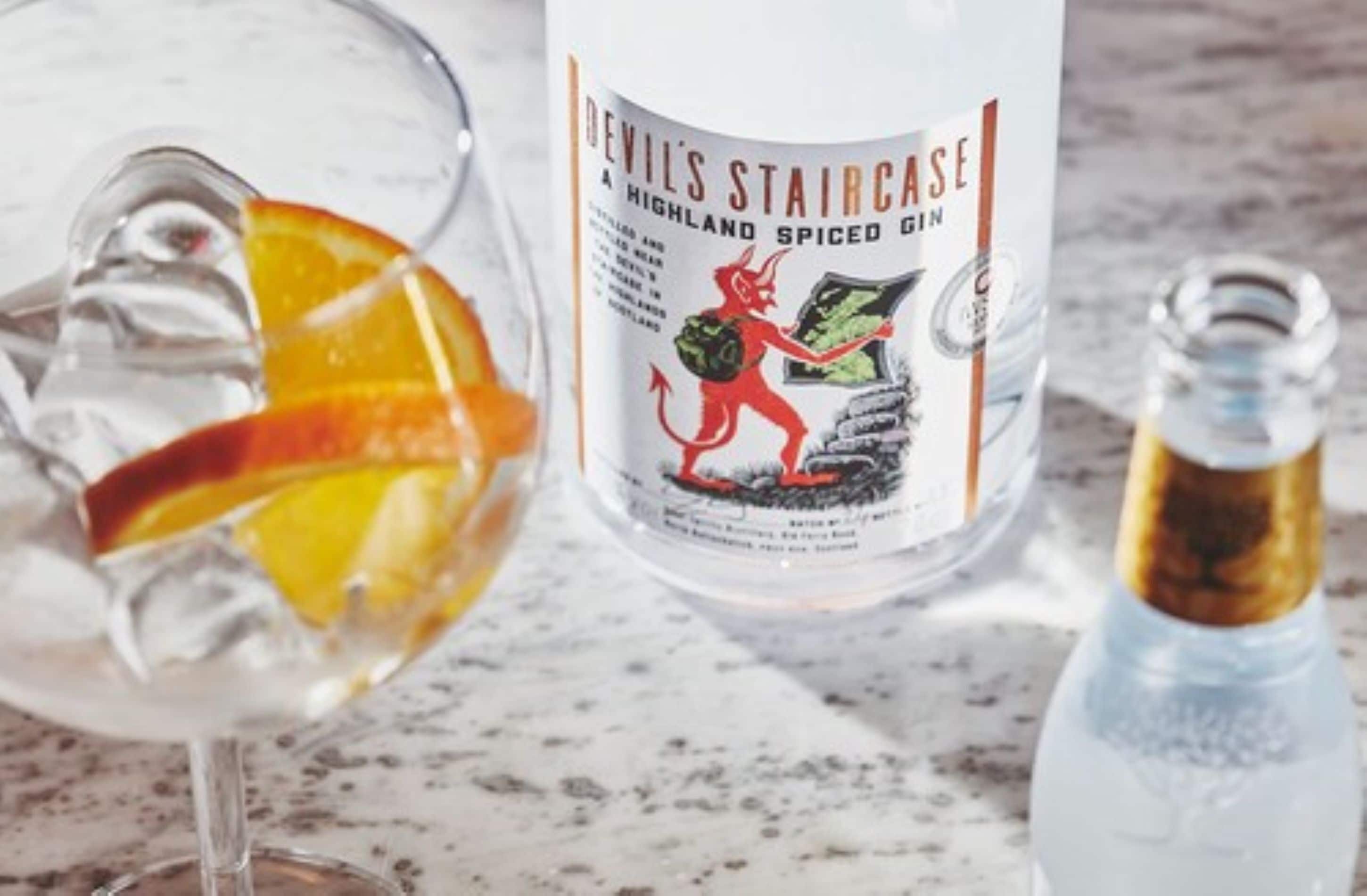 A bottle of Devil's Staircase Highland Spiced Gin, a glass with ice and an orange slice, and a bottle of clear liquid.