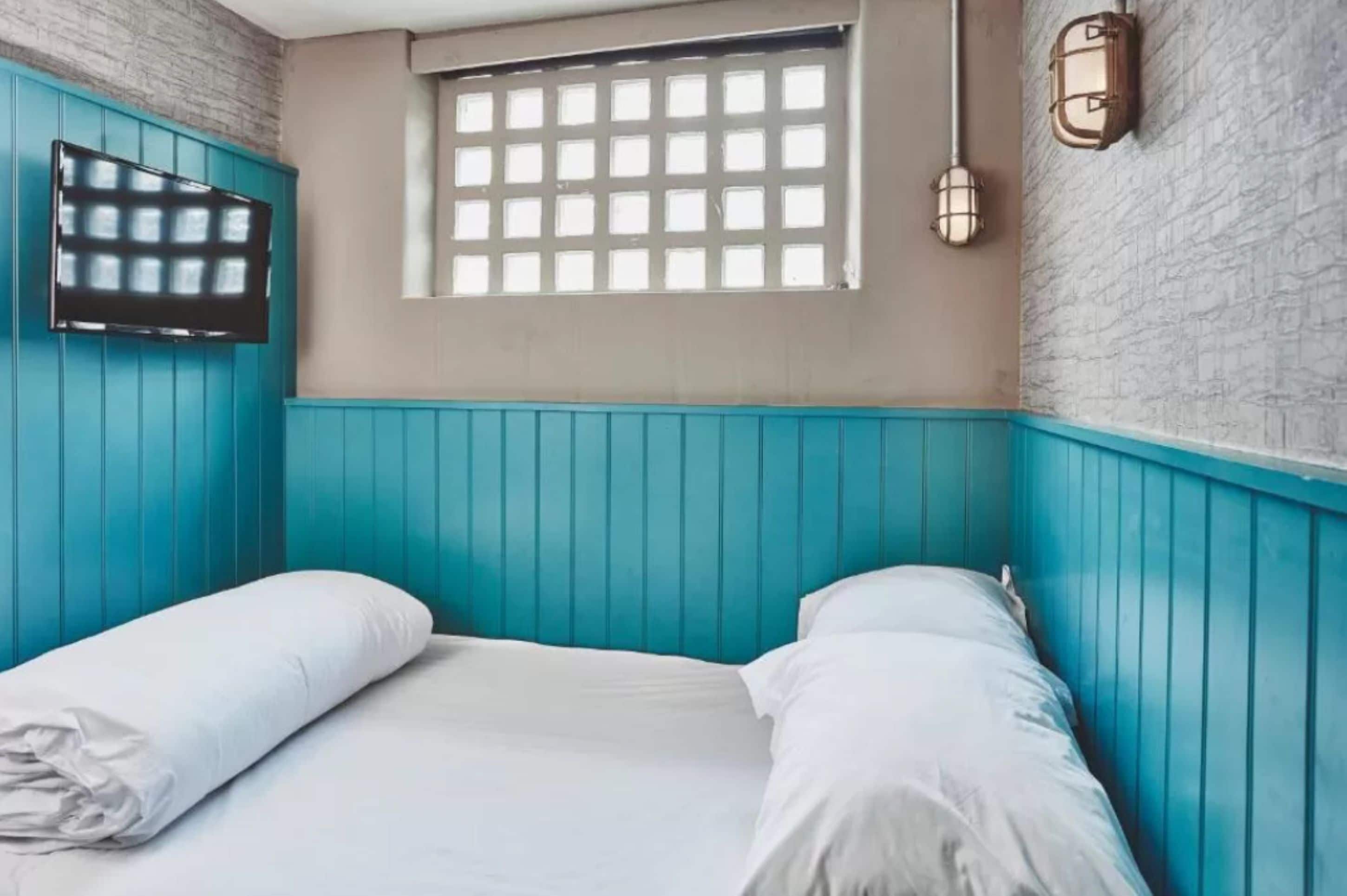 A room with teal walls, a window with a grid pattern, a flat-screen TV mounted on the wall, and two bedside lamps.