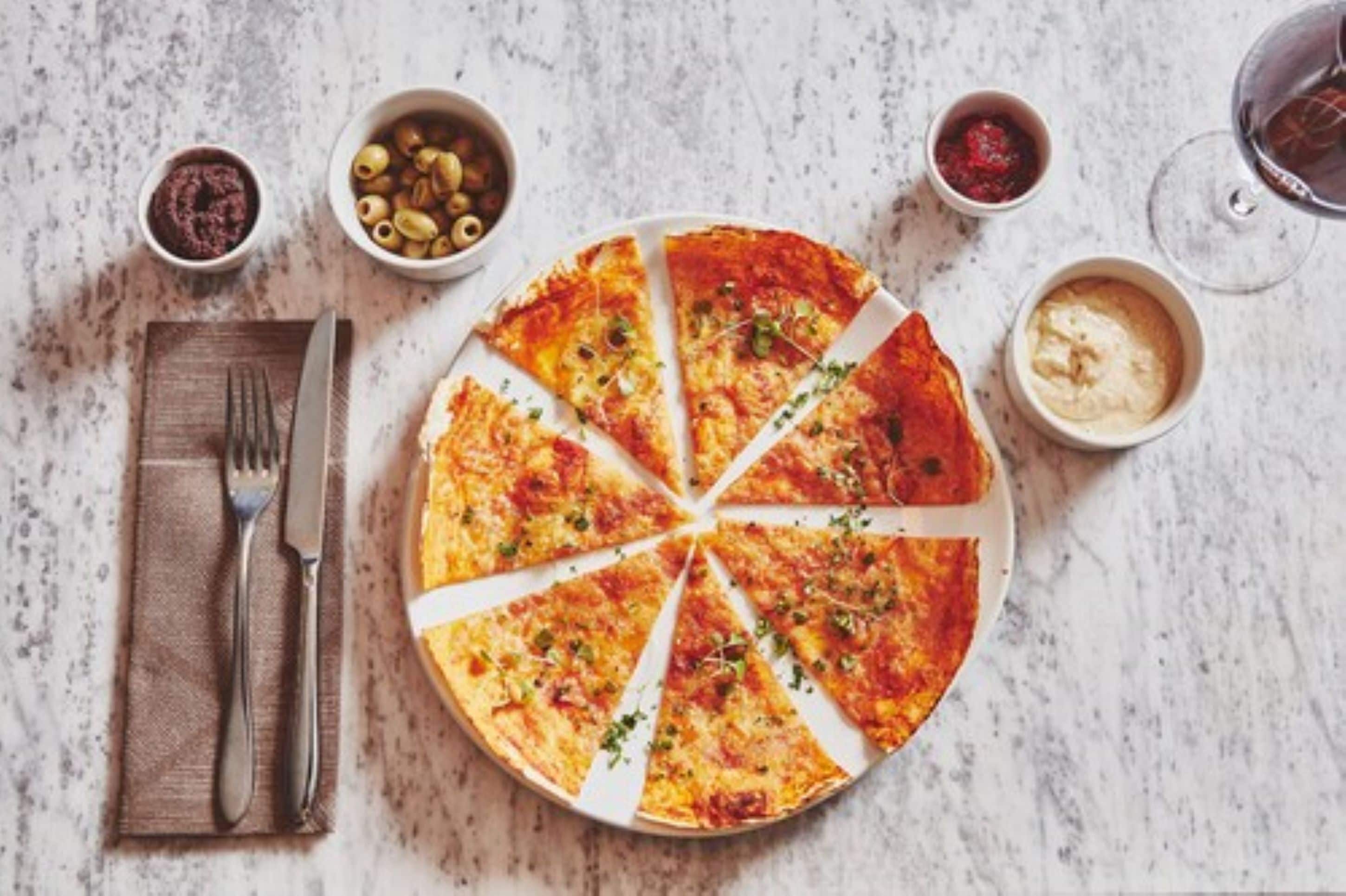 Una pizza con ocho porciones sobre un plato blanco, acompañada de recipientes pequeños con aceitunas, salsa marinara y una salsa cremosa.