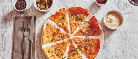 Una pizza con ocho porciones sobre un plato blanco, acompañada de recipientes pequeños con aceitunas, salsa marinara y una salsa cremosa.