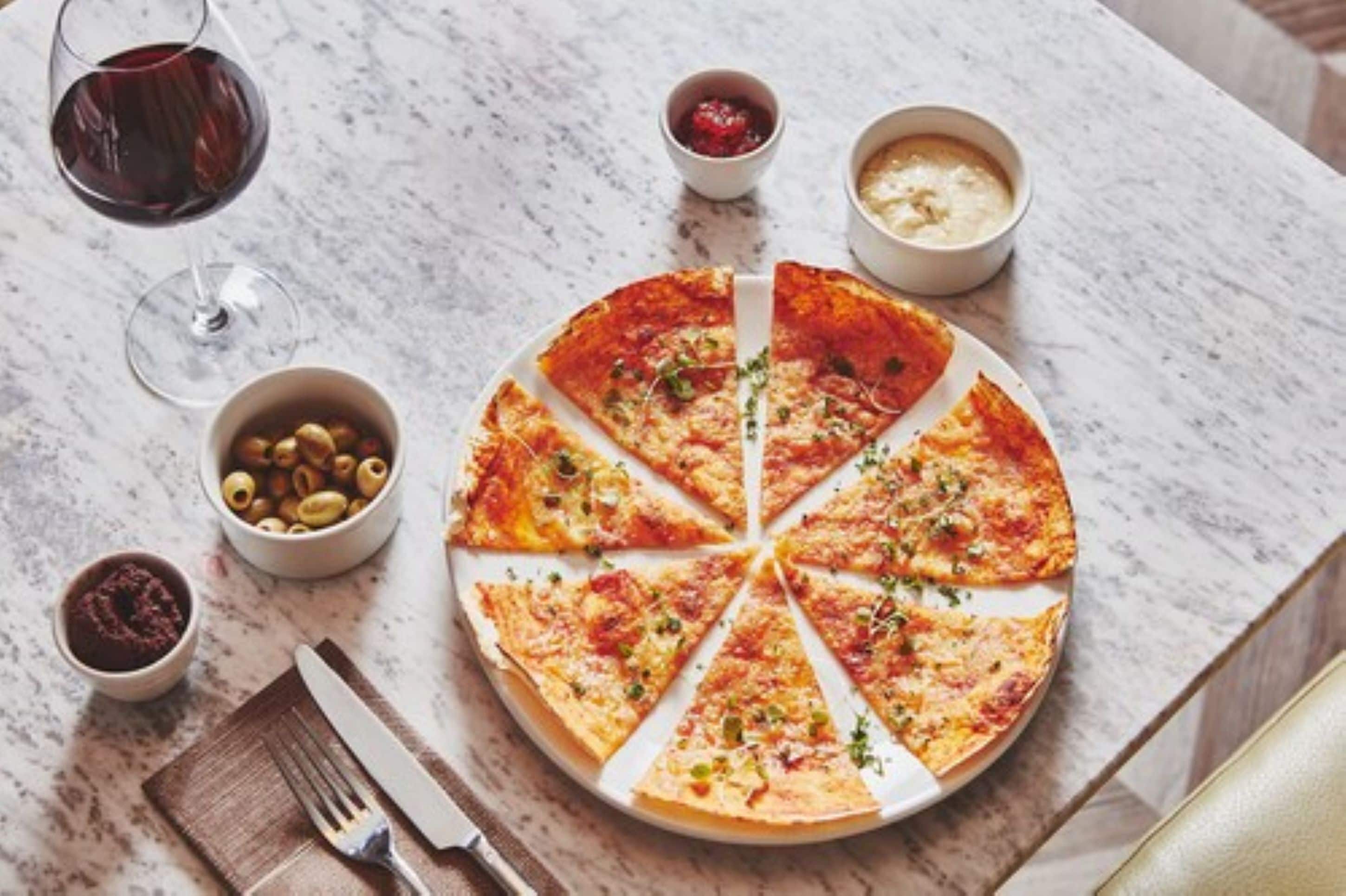 A pizza on a marble table with a fork and knife, a glass of red wine, and small bowls of olives and sauces.