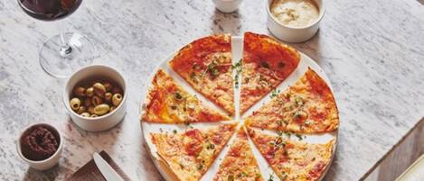 A pizza on a marble table with a fork and knife, a glass of red wine, and small bowls of olives and sauces.