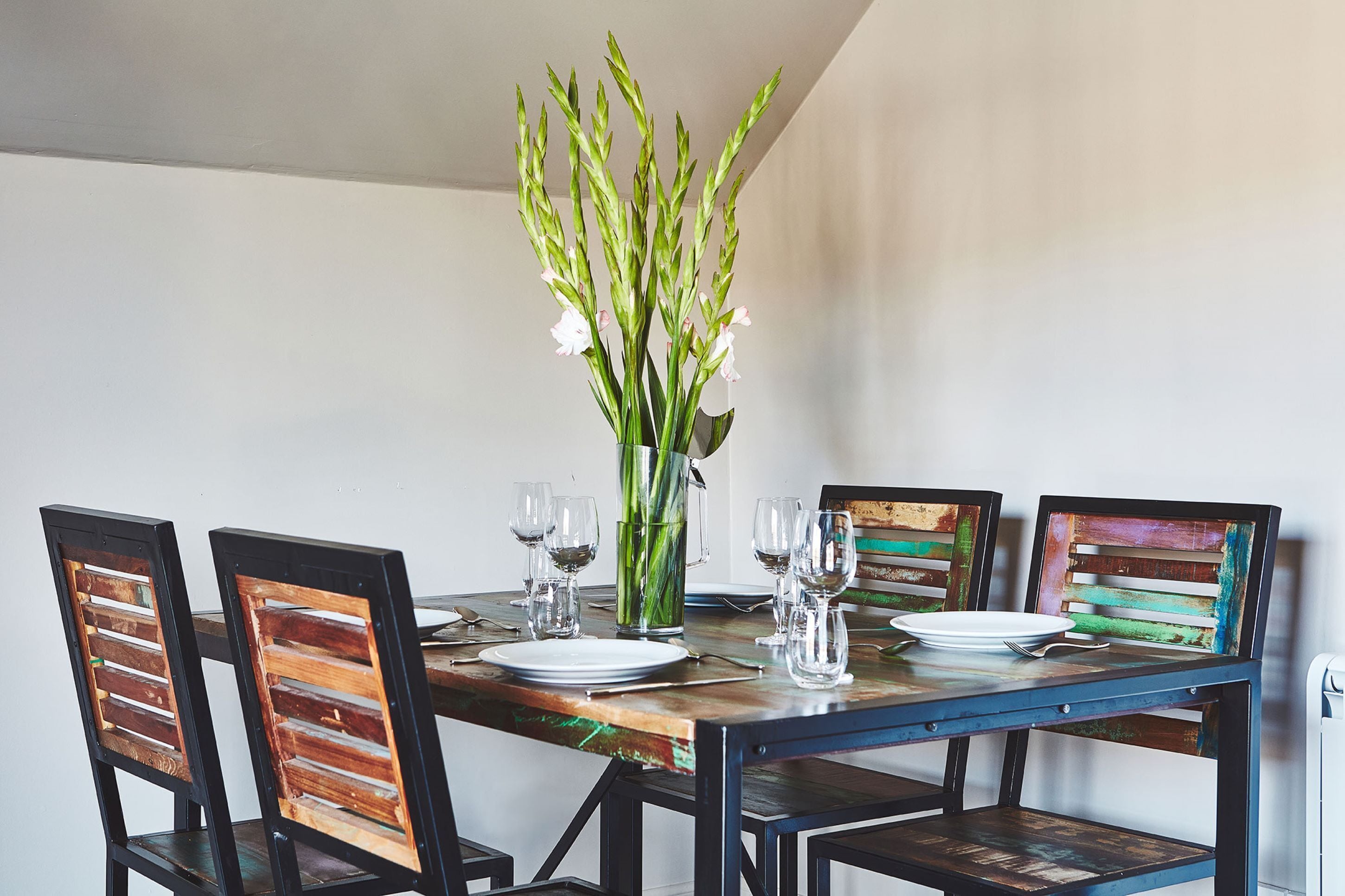 Una mesa de comedor con cuatro sillas, un jarrón con flores y copas de vino preparadas para cuatro personas.