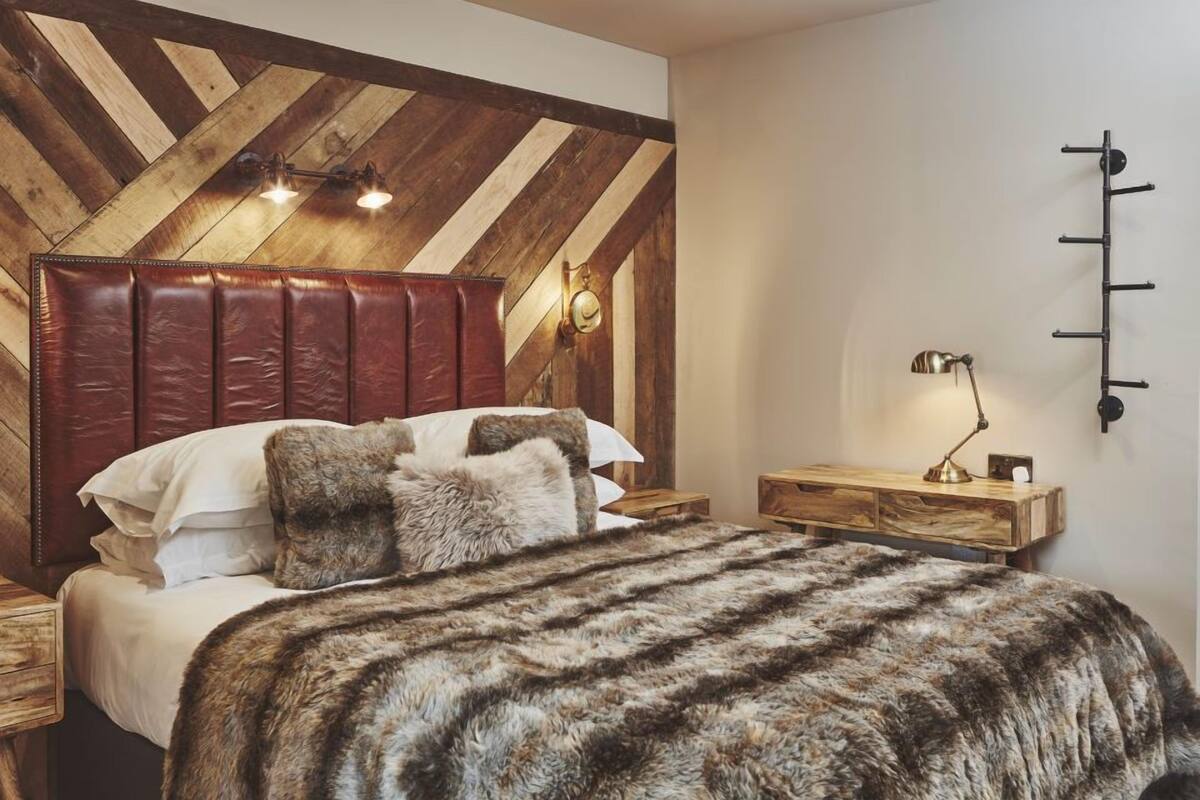 Quarto com teto de madeira, uma cama grande com manta de pele, mesa de cabeceira e luminária fixada na parede.
