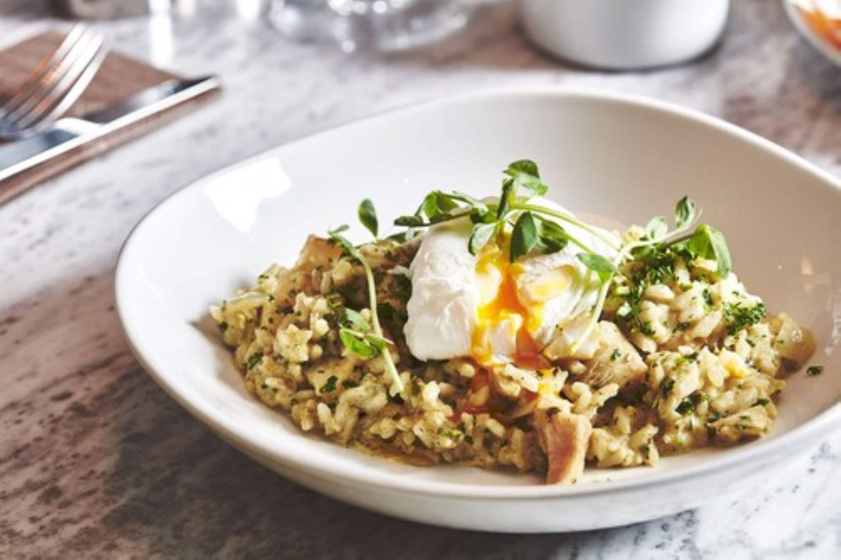 A white bowl of rice with greens and a poached egg on top, placed on a marble surface.
