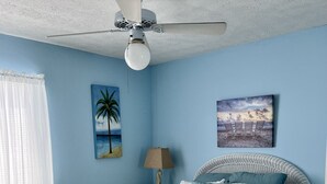 A bedroom with a bed, a ceiling fan, a nightstand, a lamp, and two wall art pieces. - Knee Deep in the Water Somewhere... (Key Colony Beach)