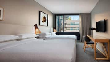 Habitación de hotel con dos camas, un escritorio, una silla, un televisor y un amplio ventanal con vistas a edificios.