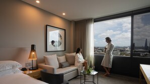 A hotel room with a bed, a sofa, a small table with a plant, a lamp, and a large window with a city view. - Four Points By Sheraton Sydney, Central Park (Chippendale)