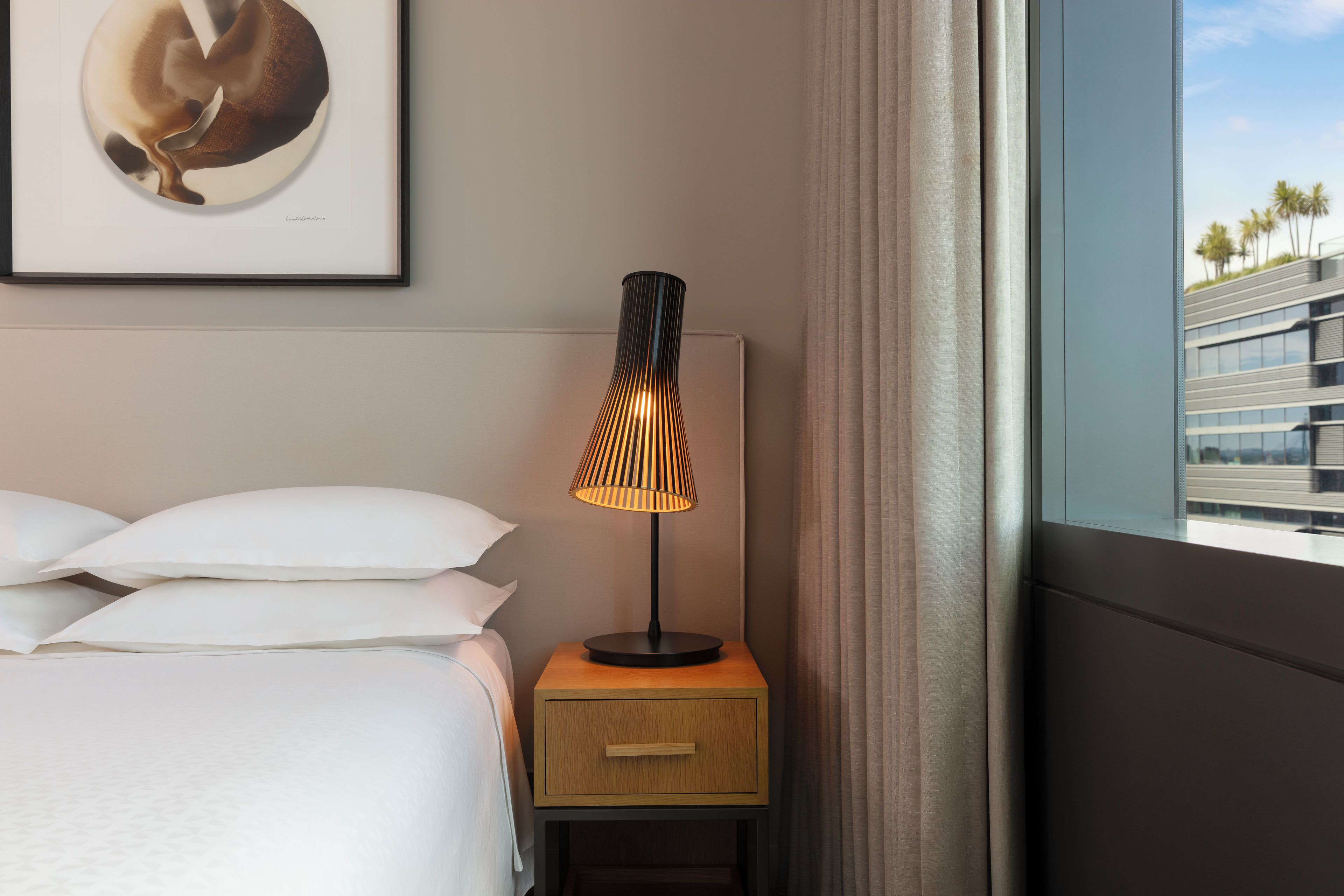 A hotel room with a bed, a bedside table with a lamp, a framed picture on the wall, and a view of a building through the window.