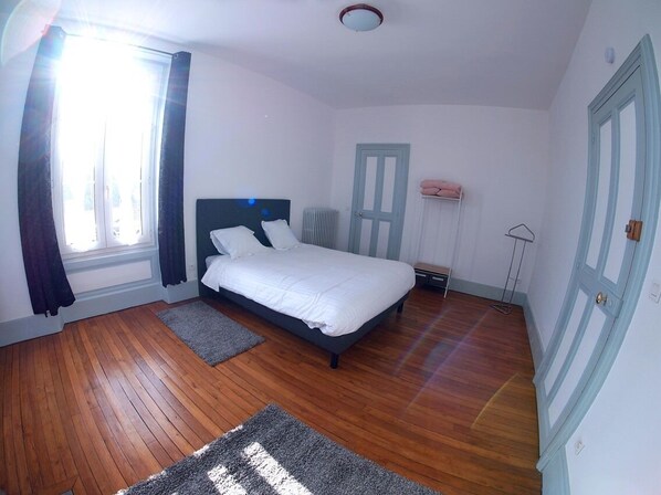 A bedroom with a bed, wooden flooring, a radiator, a door, and a window with curtains. - La Maison D'hôtes de Toucy (Toucy)