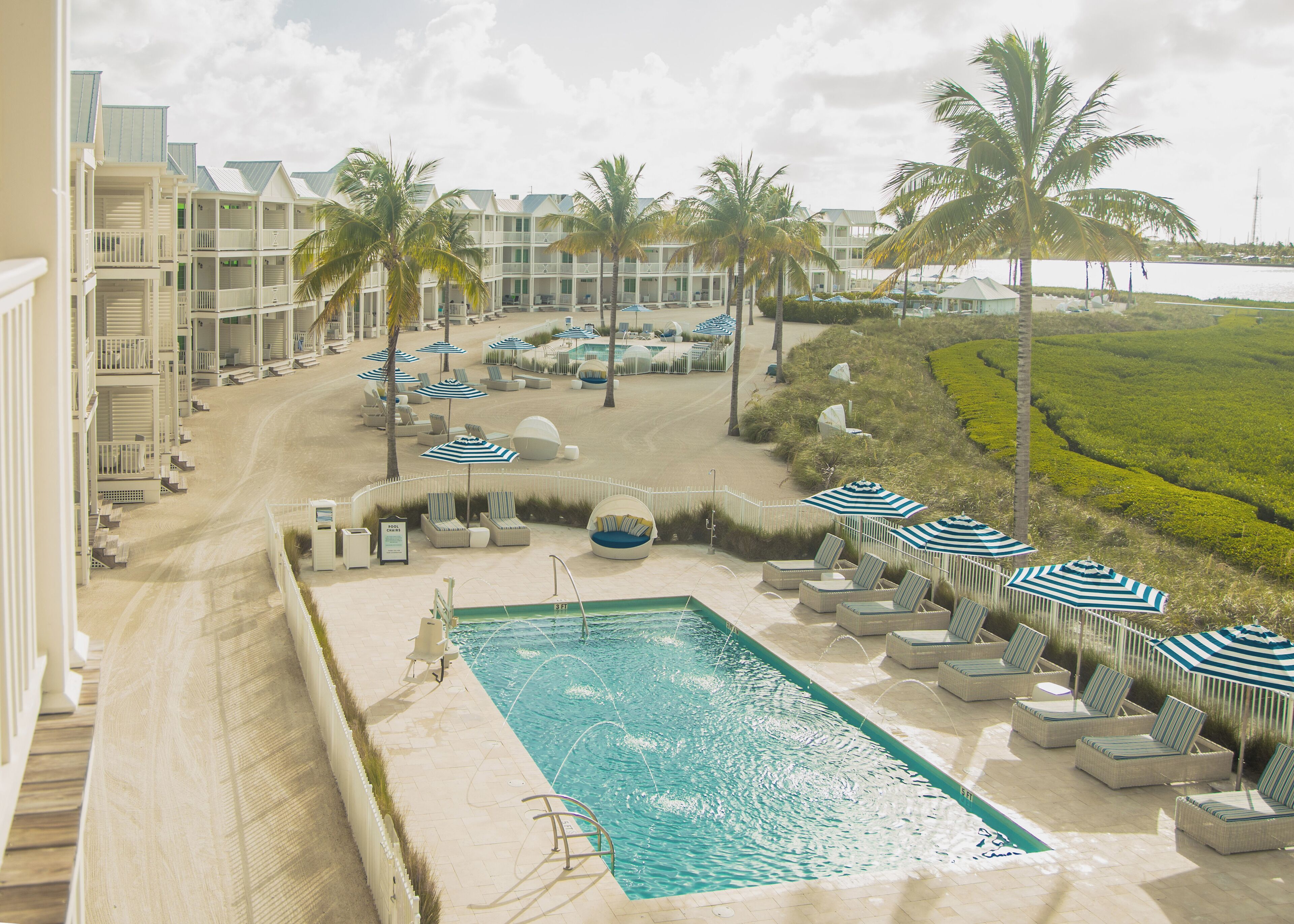 A resort pool area with lounge chairs and umbrellas, surrounded by palm trees and a view of the ocean.
