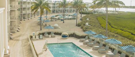 A resort pool area with lounge chairs and umbrellas, surrounded by palm trees and a view of the ocean.