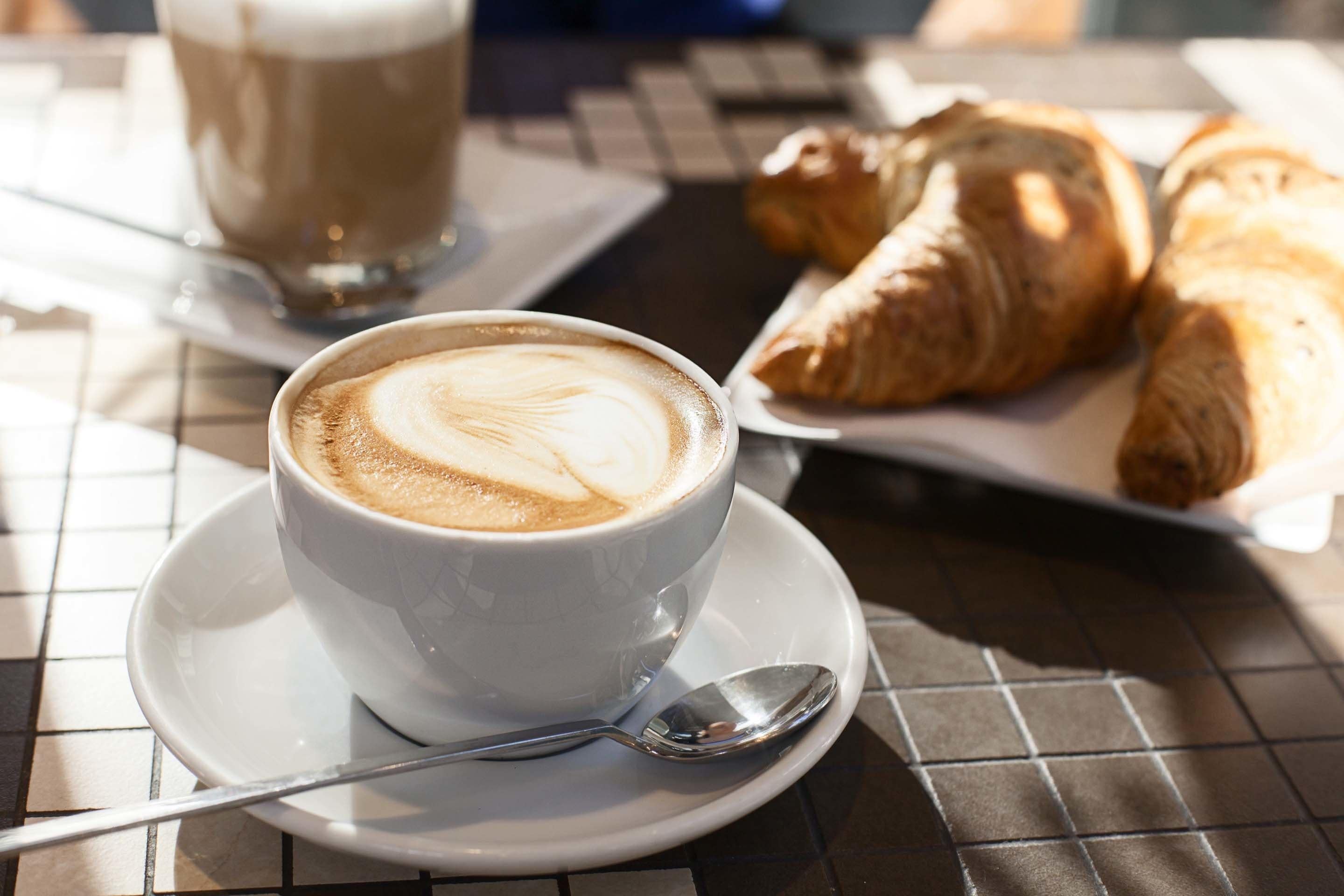 A cup of coffee with latte art on a white saucer, a spoon, and a plate with croissants.