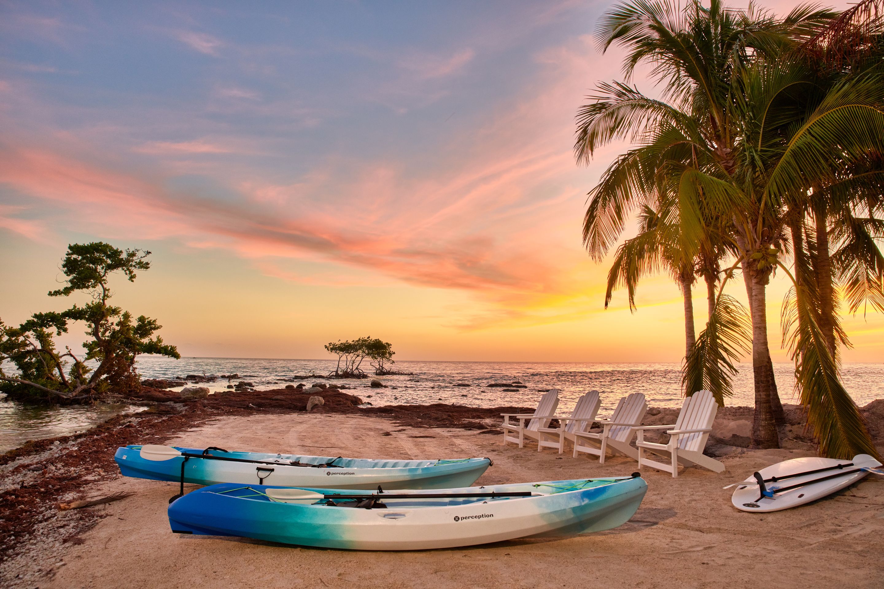 Des kayaks et des chaises sur une plage bordée de palmiers, au coucher du soleil.