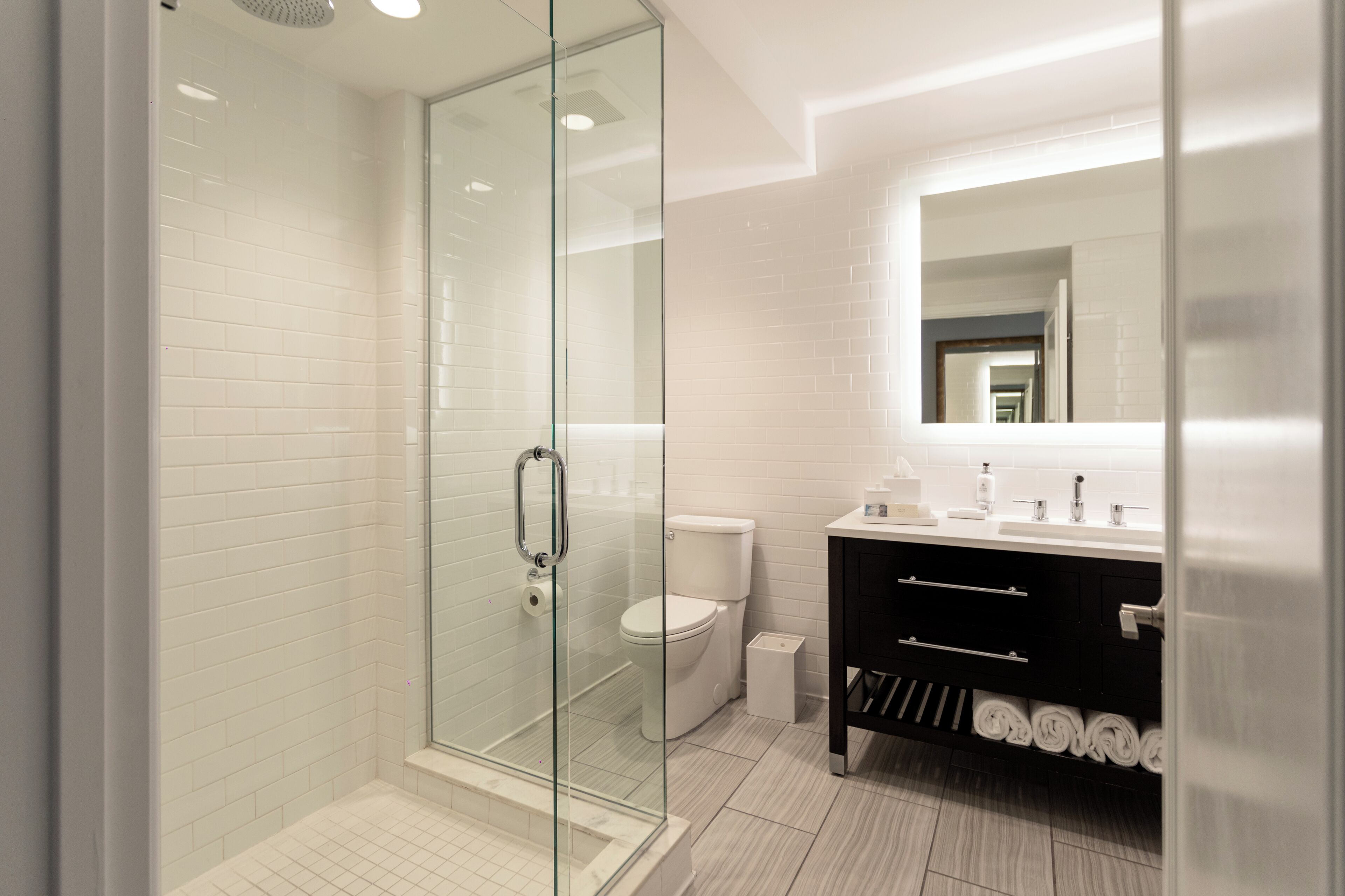 A modern bathroom with a glass-enclosed shower, white tiles, a black vanity with a mirror, and a white toilet.