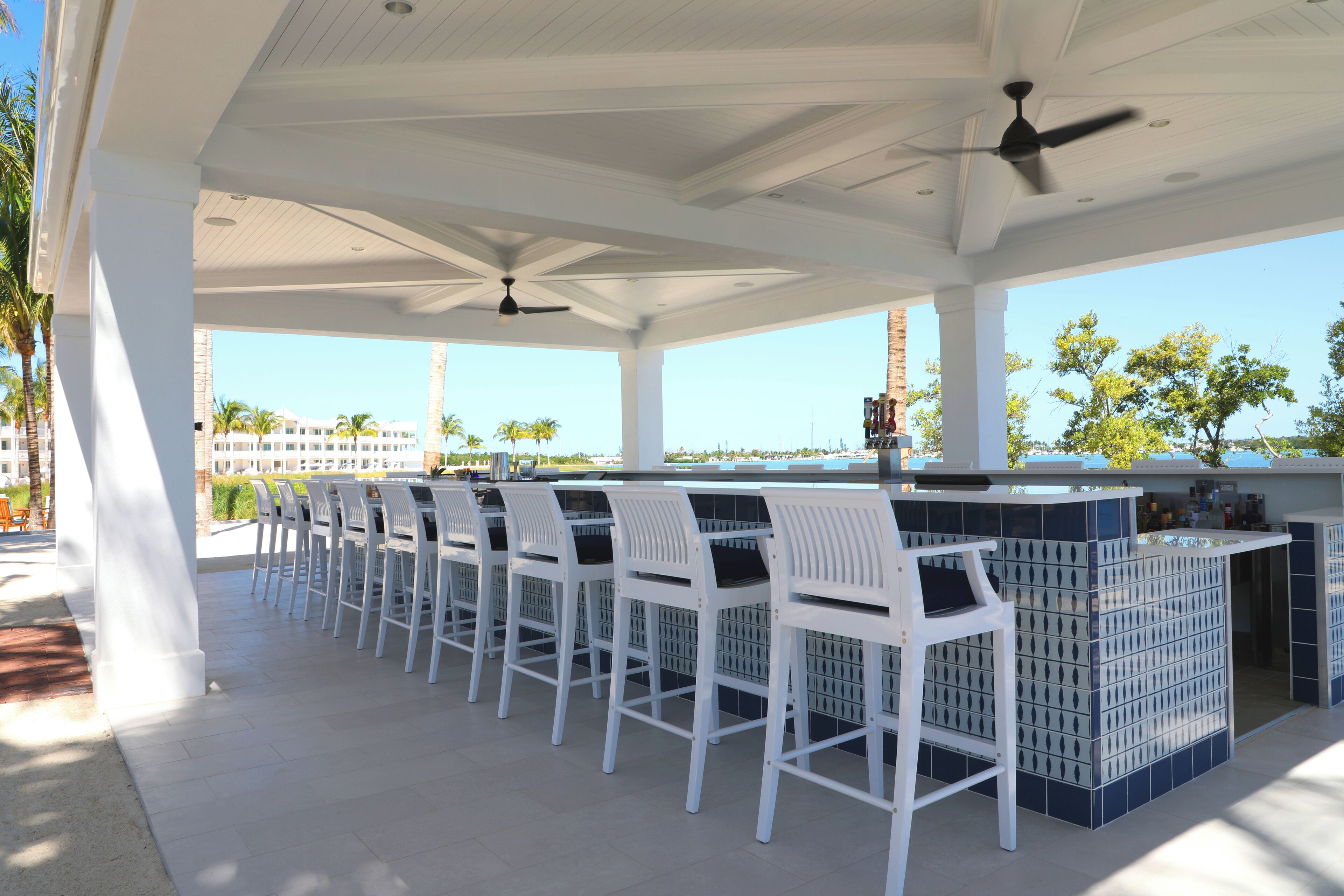 An outdoor bar area with white chairs and a counter, featuring a clear view of the ocean and palm trees.