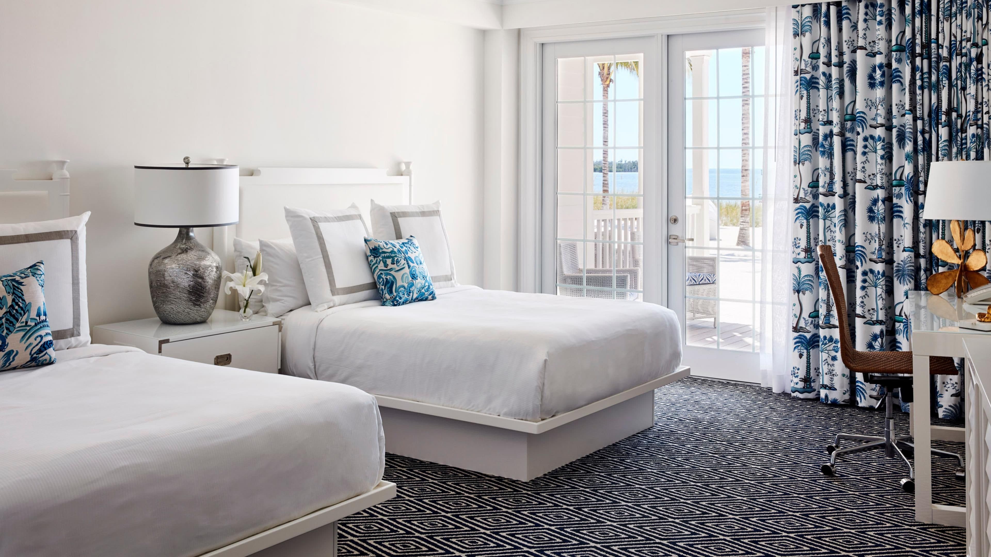 Quarto de hotel com duas camas, uma escrivaninha, uma cadeira, um abajur e vista para o exterior através de uma porta de vidro.
