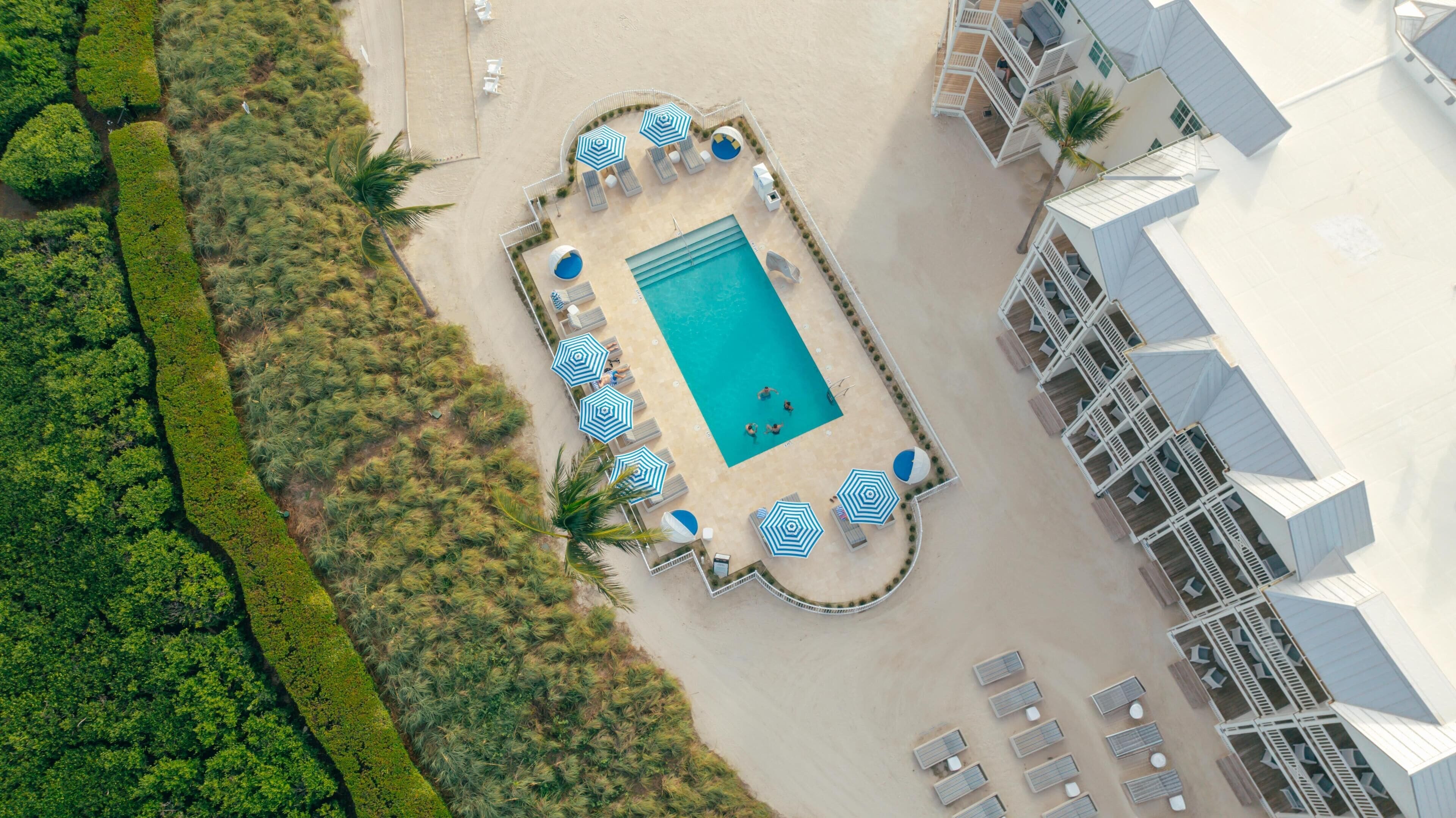 Luchtbeeld van een resort met een centraal zwembad, omringd door ligbedden en parasols, grenzend aan een gebouw met meerdere balkons.
