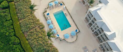 Vista aérea de un resort con piscina central, rodeada de sillas de descanso y sombrillas, y adyacente a un edificio con varios balcones.
