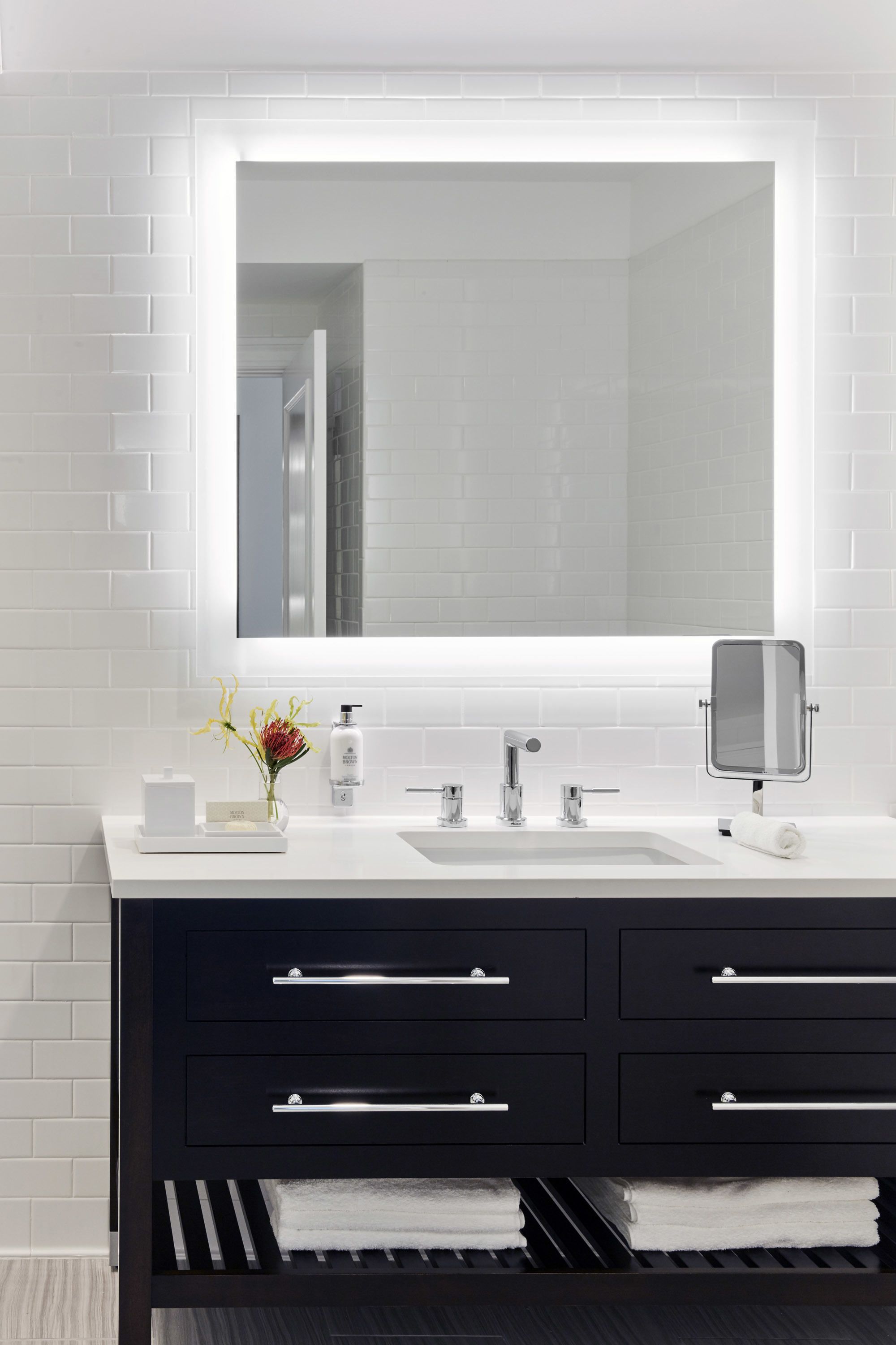 A modern bathroom with a white tiled wall, a large mirror, a white sink, and a black vanity with drawers.