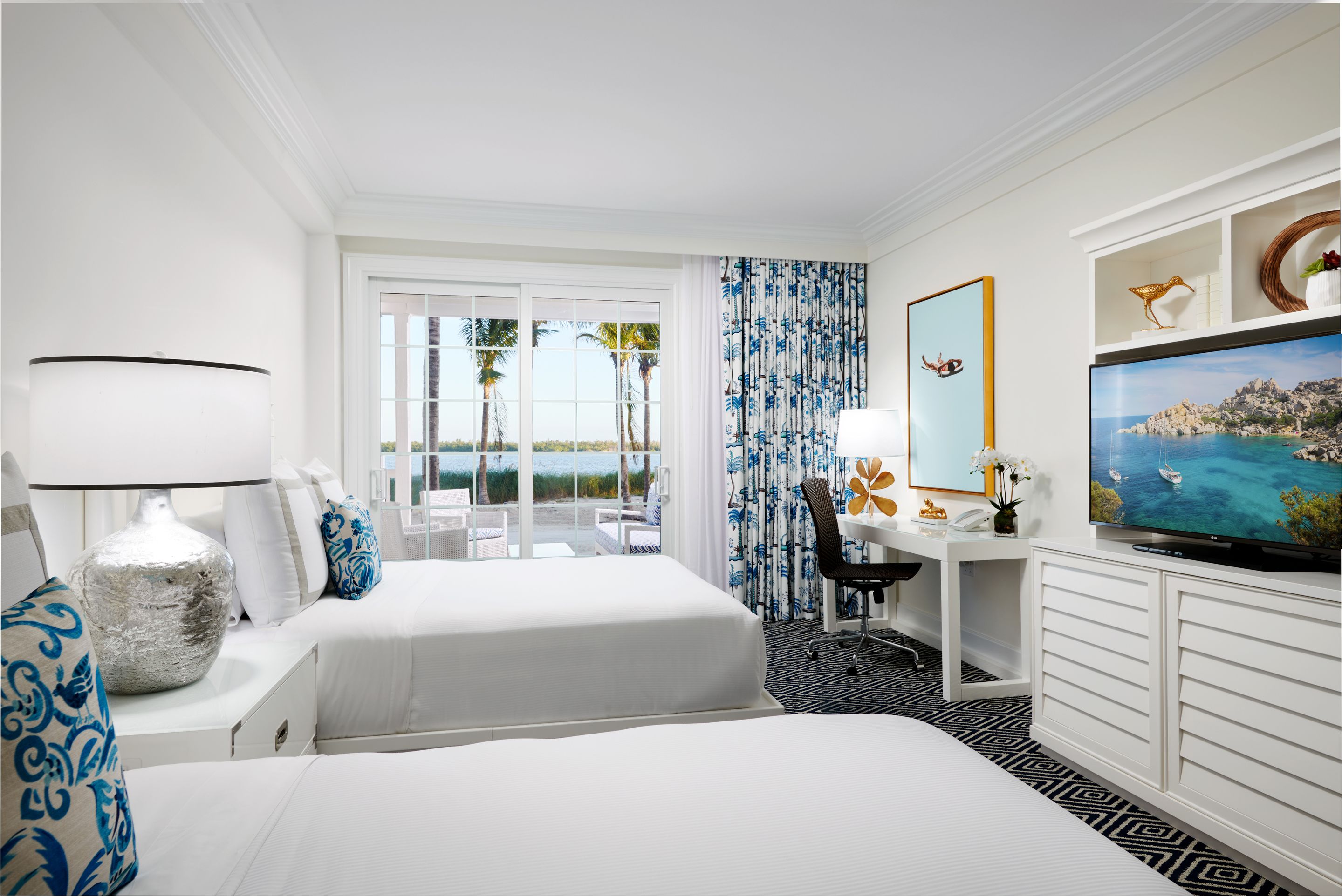 A hotel room with two beds, a desk with a chair, a television, and a view of the beach.