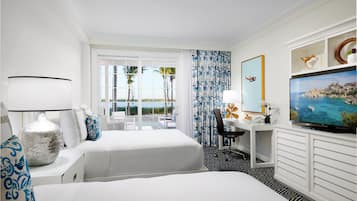 A hotel room with two beds, a desk with a chair, a television, and a view of the beach.