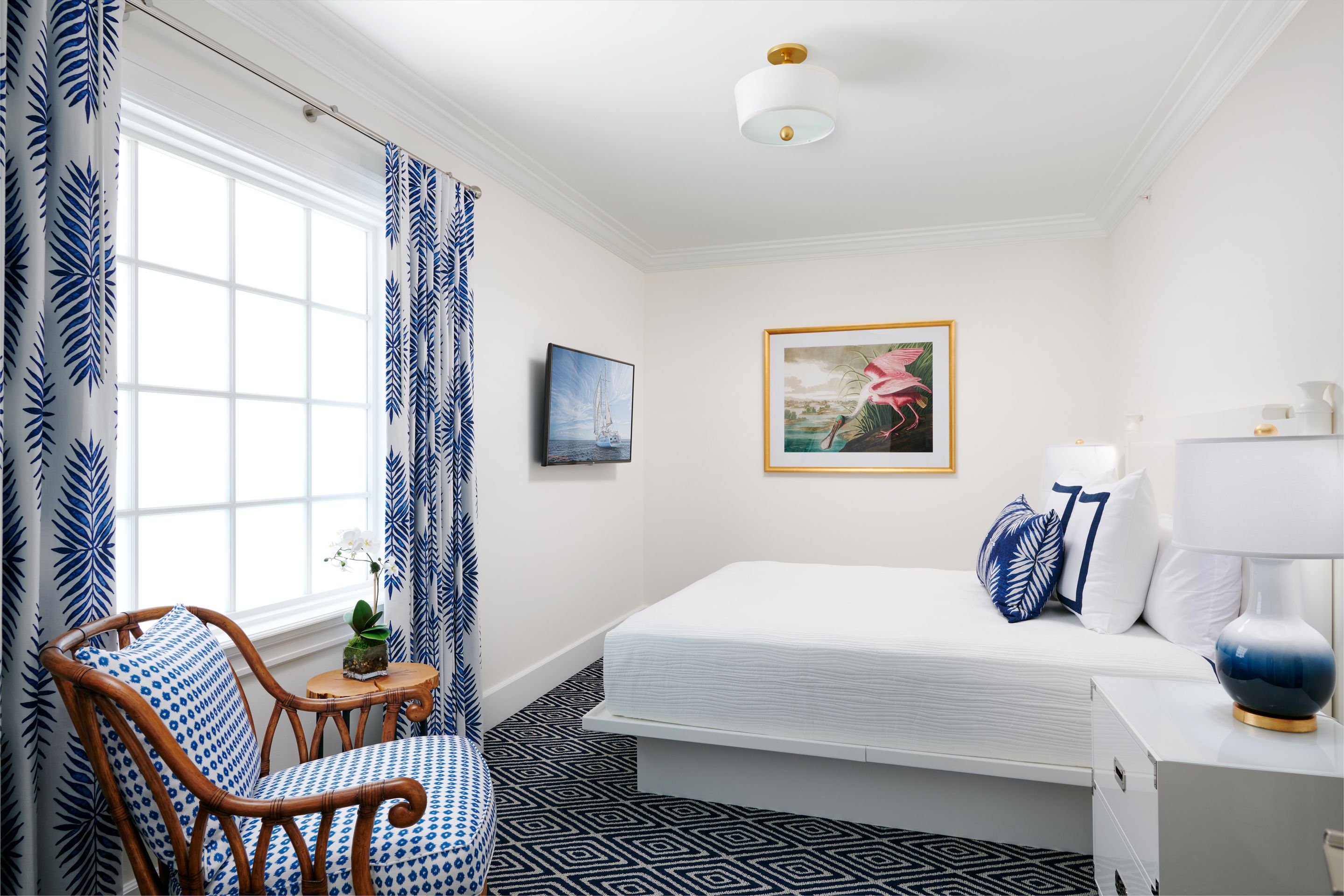 A hotel room with a bed, a chair, a small table with a plant, a framed picture on the wall, and a lamp on the bedside table.