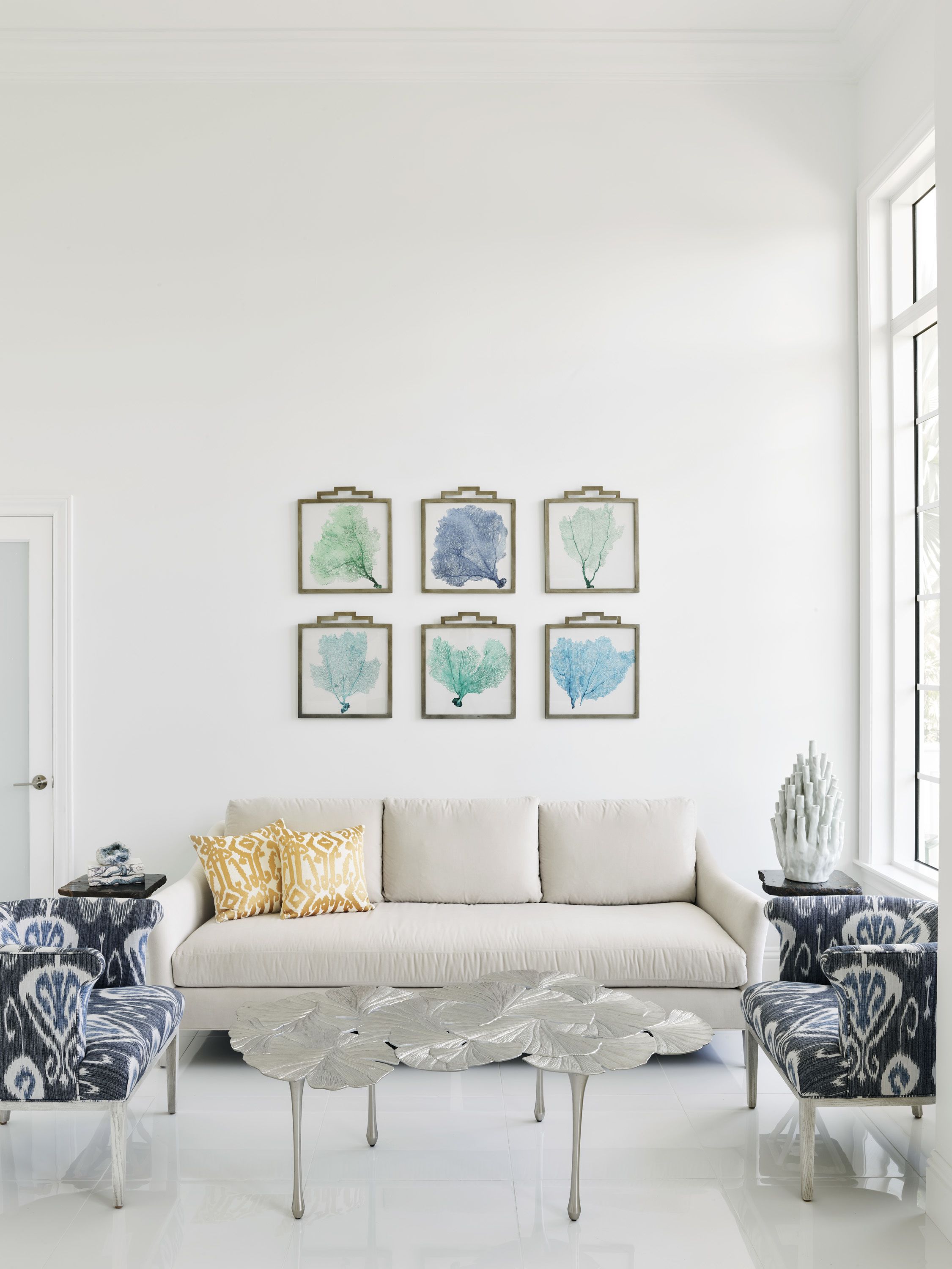 A modern living room with a white sofa, patterned armchairs, a glass coffee table, and framed artwork on the wall.