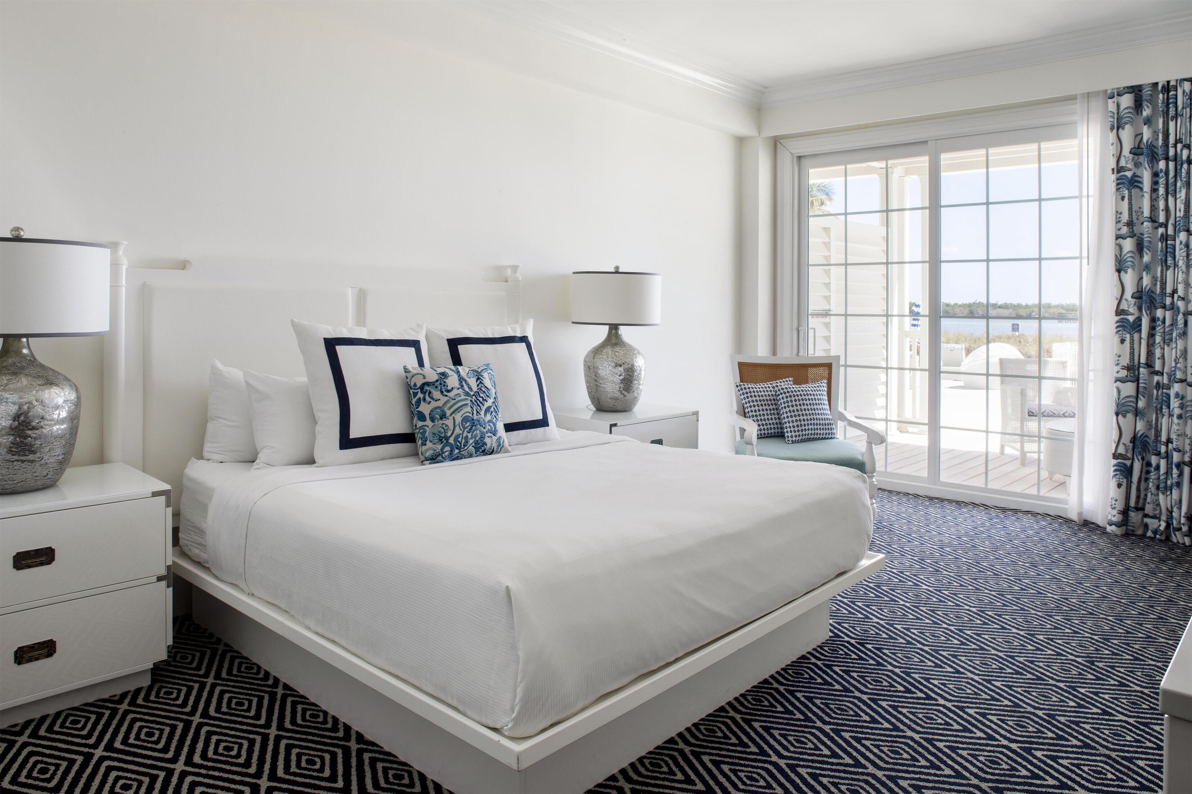 Habitación de hotel con una cama grande, dos mesitas de noche, una silla y vista al mar a través de amplios ventanales.