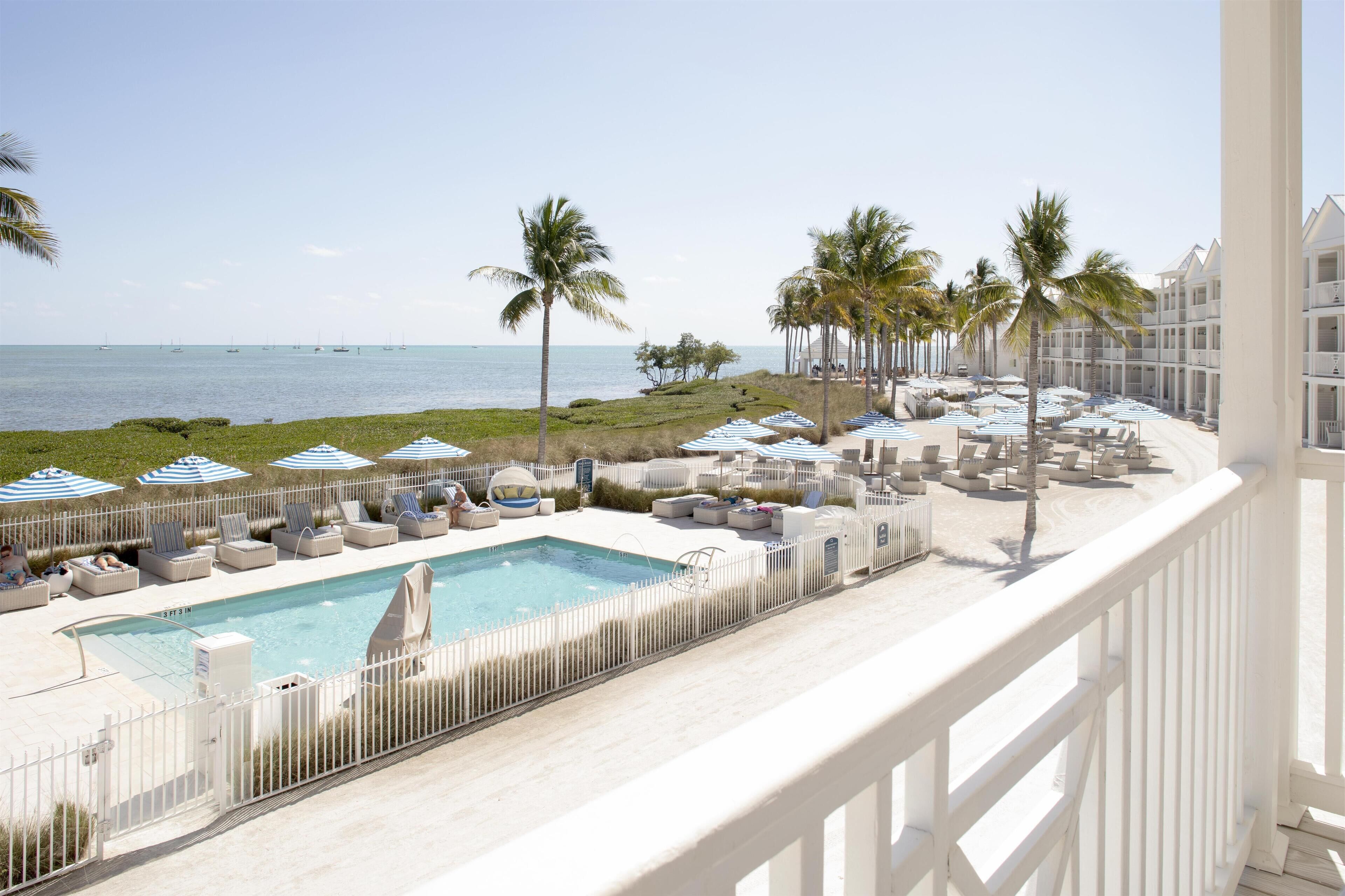 A pool area with lounge chairs and umbrellas, a clear view of the ocean, and a white fence surrounding the pool.