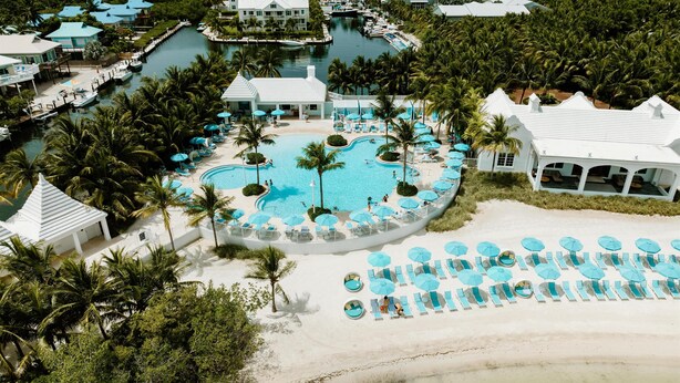 Vue aérienne d’un complexe hôtelier doté d’une piscine centrale, de plusieurs bâtiments et d’une plage aménagée avec des transats.