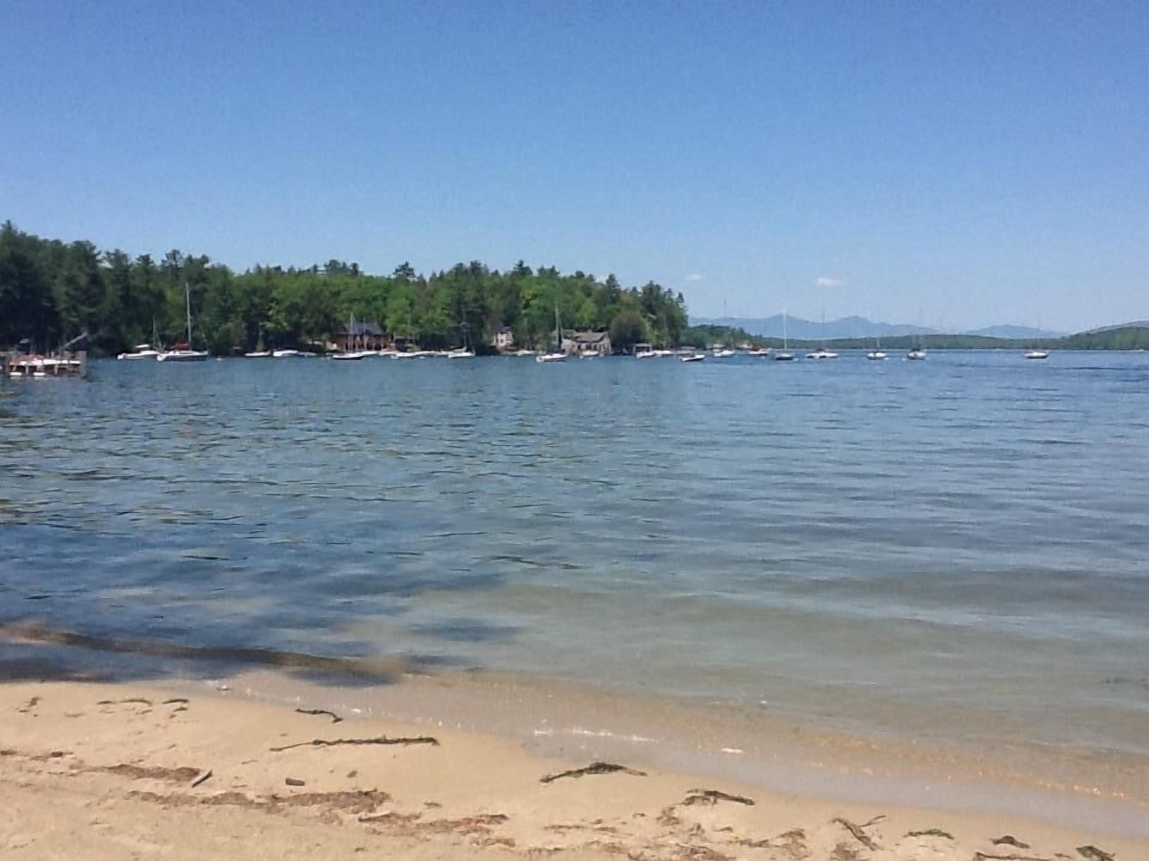 A beach with clear water, boats, and a forested area in the background.