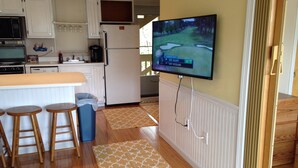 Una cocina con televisor montado en la pared mostrando un partido de golf, taburetes de bar de madera, un cesto de basura y alfombras estampadas.