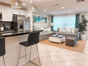A modern kitchen with a refrigerator, microwave, and toaster. There is a dining area with a bar stool and a coffee table. The living room has a sofa set and a large window with curtains.