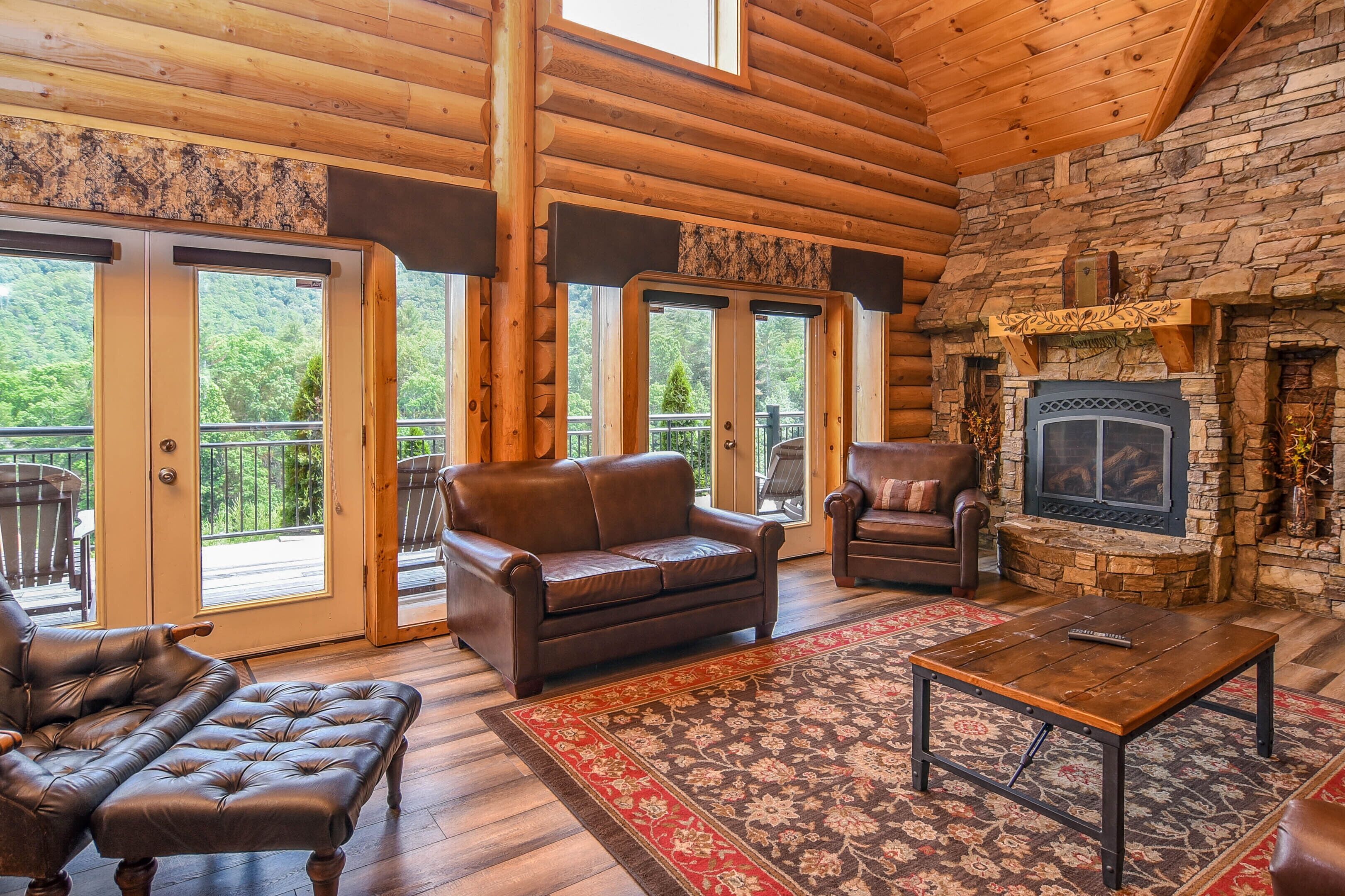 A log cabin interior with a fireplace, leather furniture, and large windows offering a view of the forest.
