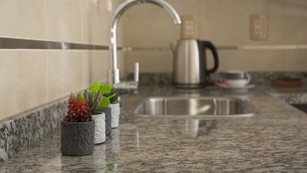Una cocina con encimeras de granito, un lavamanos de acero inoxidable, una tetera y pequeñas macetas.