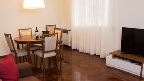 Una sala de estar con una mesa de comedor de madera, cuatro sillas, un televisor de pantalla plana sobre un soporte de madera y un cojín rojo en un sofá.