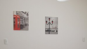 Una habitación con un sofá marrón, dos cojines rojos y dos obras de arte en la pared que representan escenas urbanas.