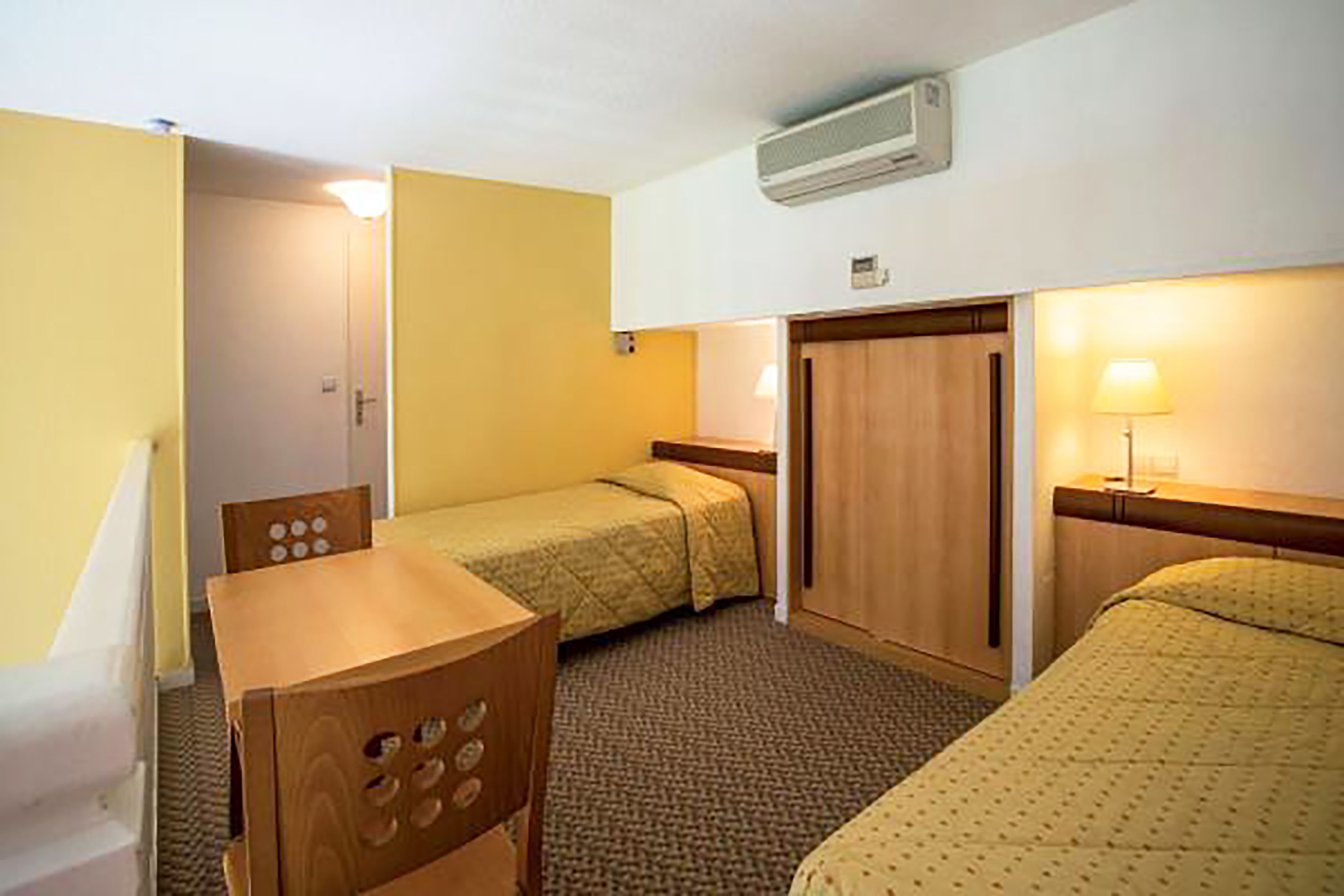 Quarto de hotel com duas camas, uma escrivaninha de madeira, uma cadeira, um abajur e um aparelho de ar condicionado.