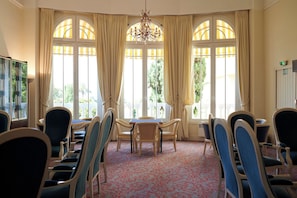 A room with a dining table, chairs, a chandelier, large windows with curtains, and a patterned carpet. - Résidence Vacances Bleues Le Méditerranée (Saint-Raphael)