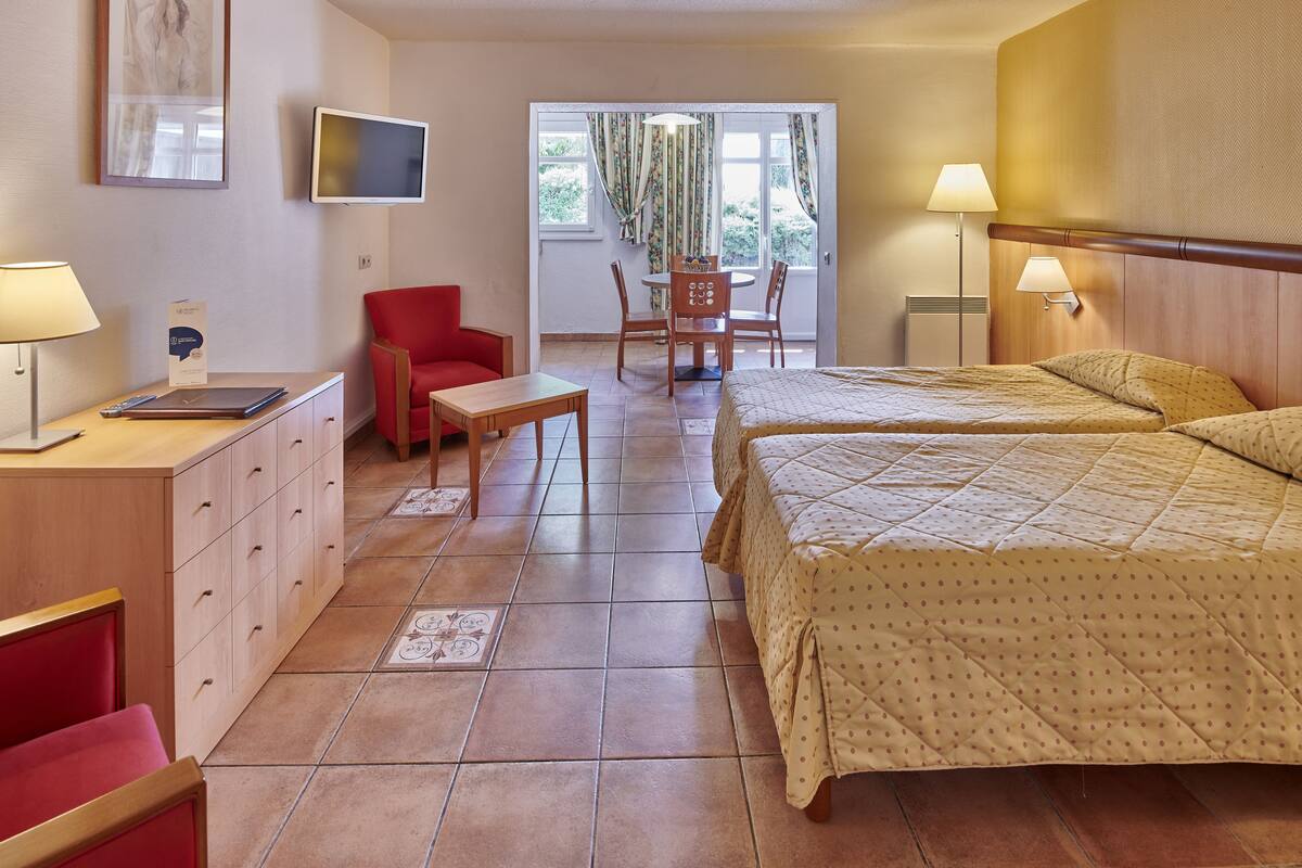 A hotel room with two beds, a desk with a chair, a red armchair, a TV, and a dining table with chairs.